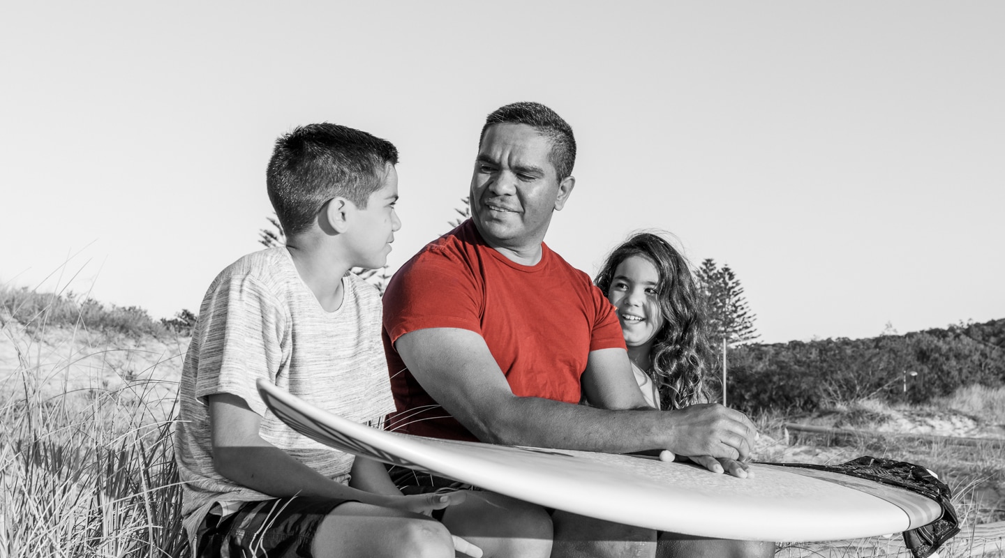 Middle aged Indigenous man explains surfing to his pre-teen children whilst at an Australian beach