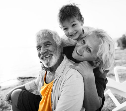 Senior couple sit on Australian beach with their grandson climbing on top of them, grinning