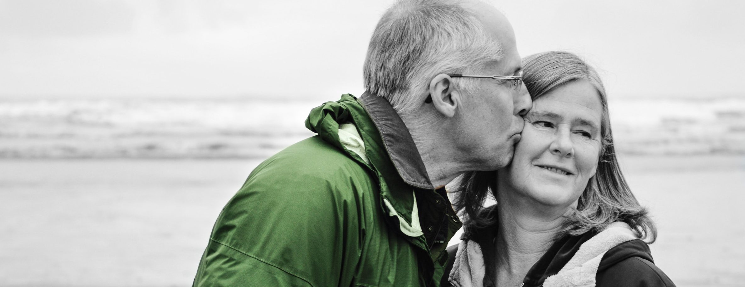 Senior couple stand on a beach as husband places a kiss on his wife's cheek