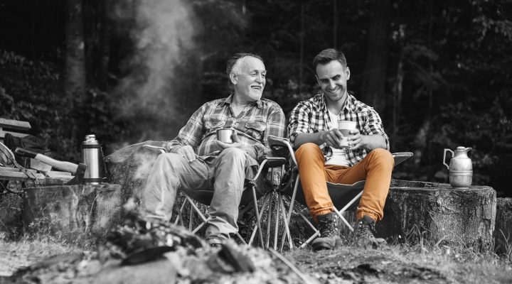 Adult son and his father sit around a campfire having a laugh over a cup of tea