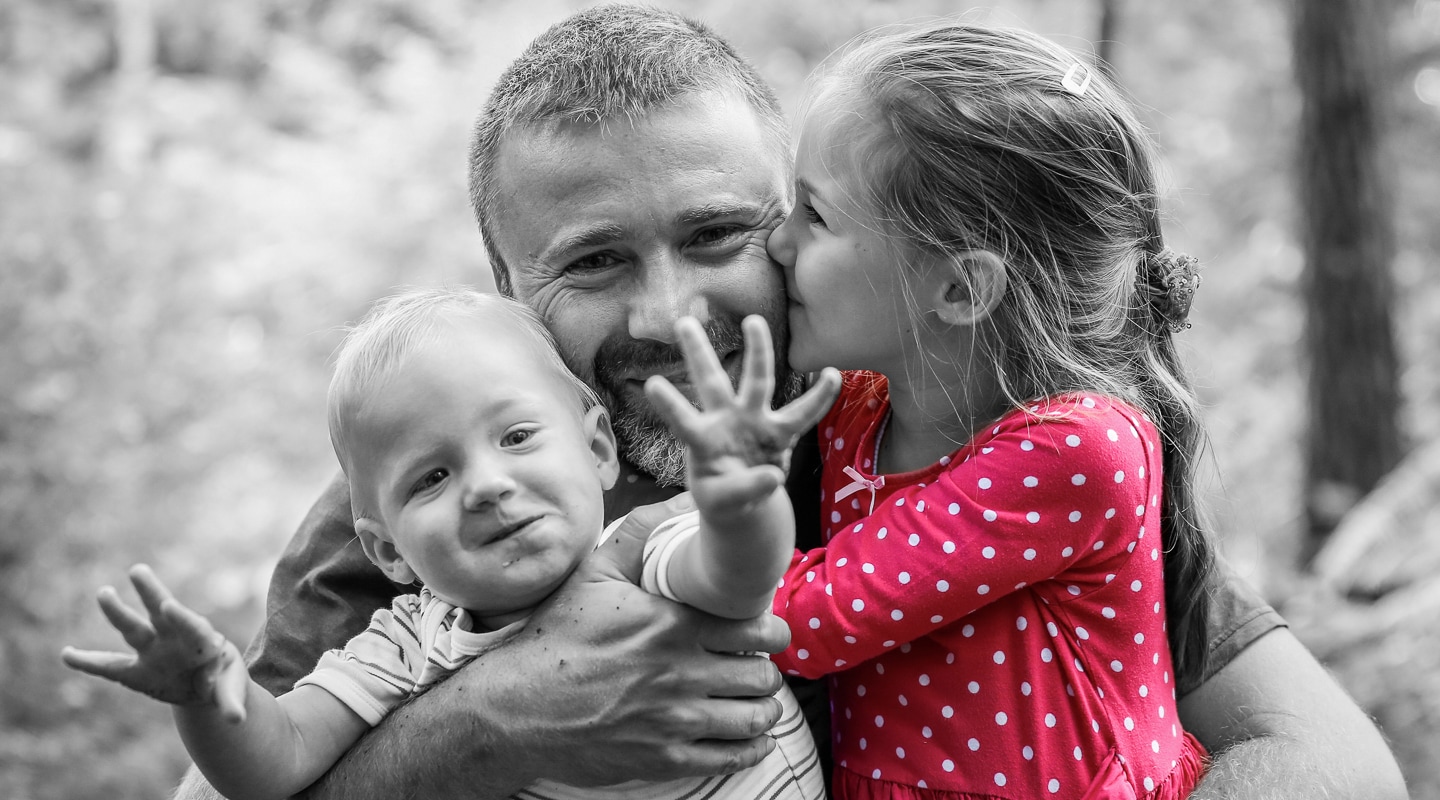 Father holds both his children as his daughter kisses his cheek and his son reaches towards his mother