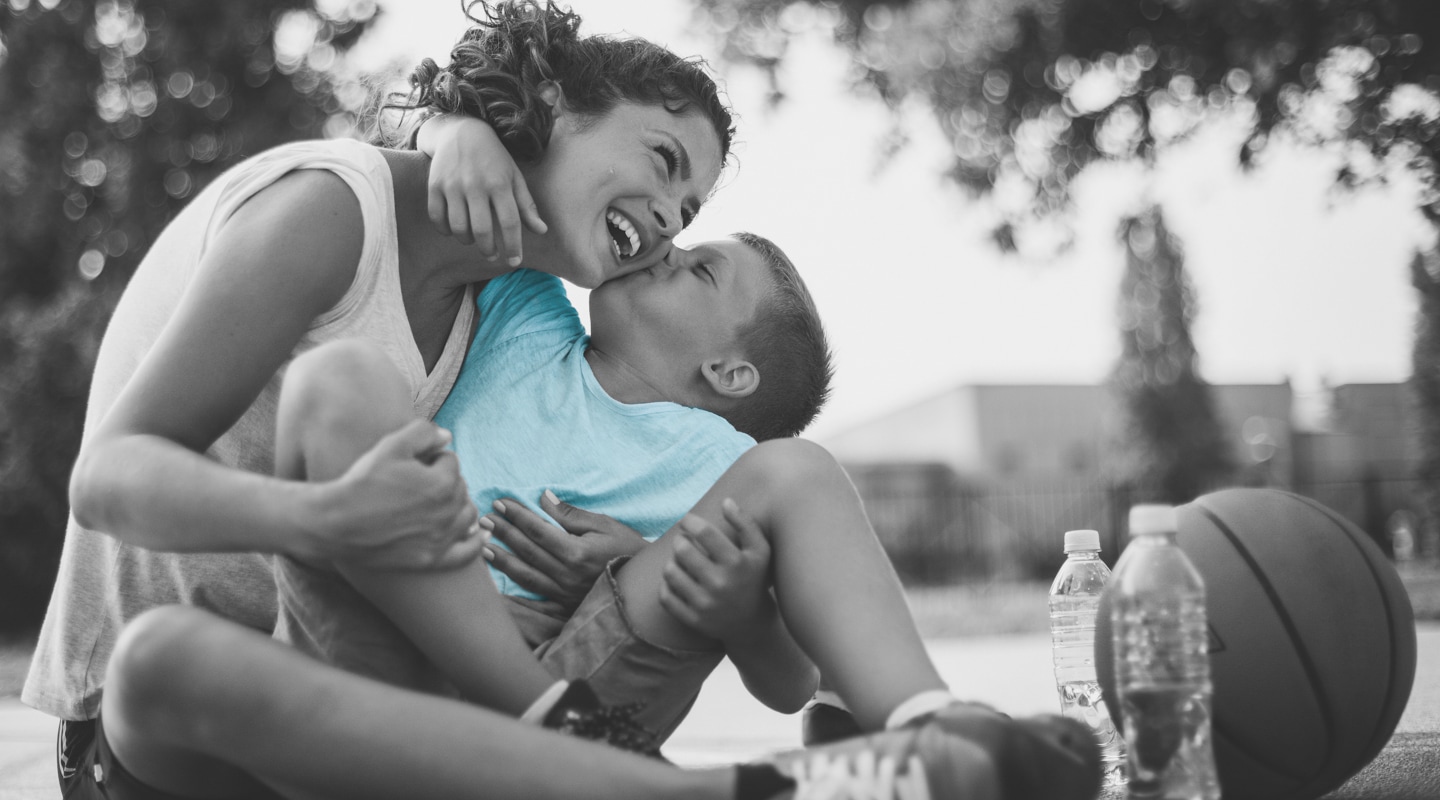 Mother laughs while her young son leans up to kiss her cheek