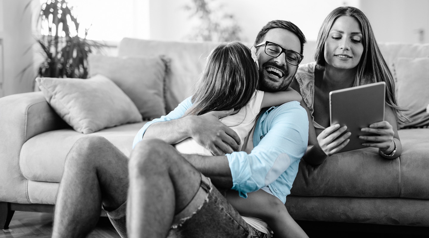 Young couple in their thirties sit in their living room while their daughter jumps into her father's arms for a hug