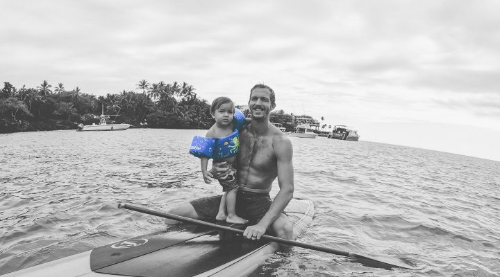Young father in his thirties holds onto his young daughter's waist as they balance on a paddle board