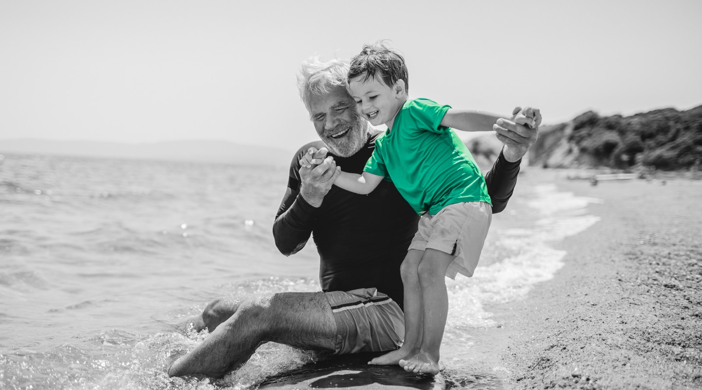 Elderly grandfather laughs with his grandson as he balances on a surfboard on an Australian beach shore