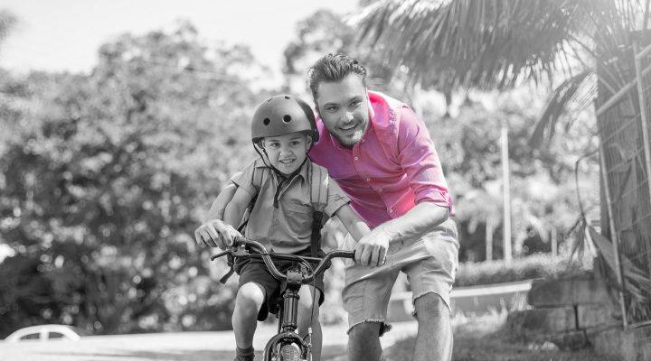 Proud father carefully steadies his young son on the bike as he goes for his first ride without training wheels