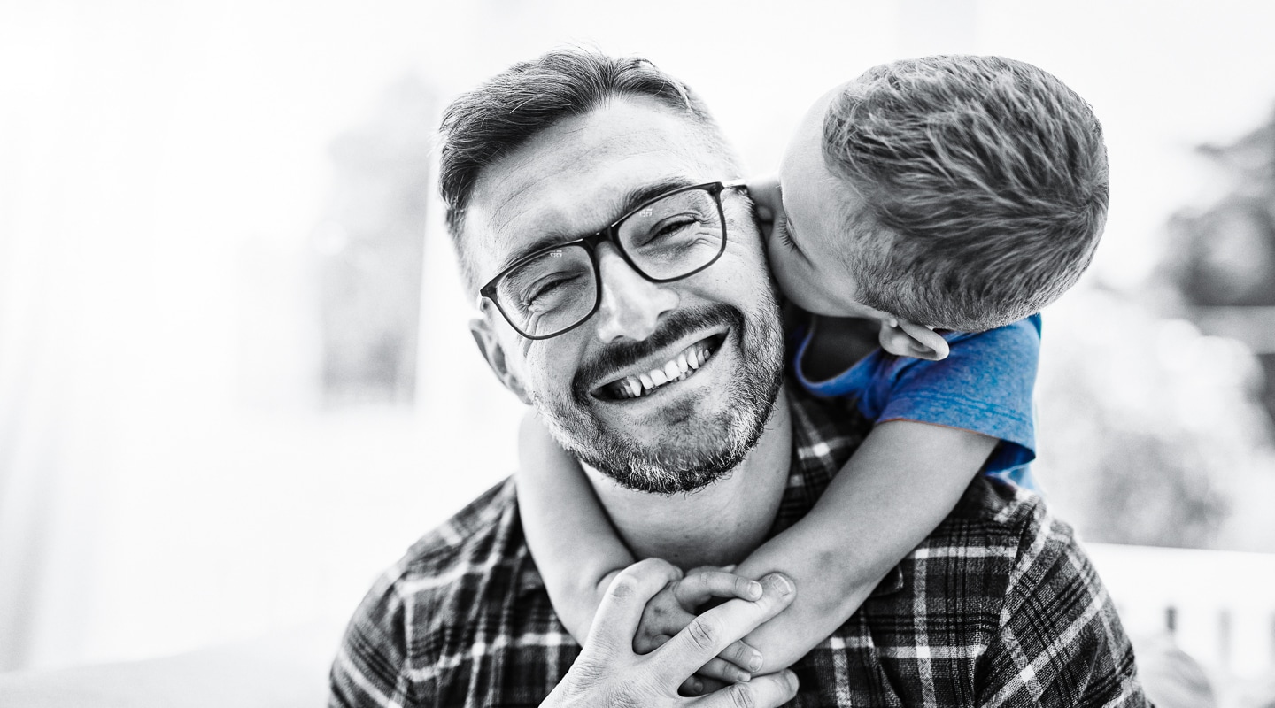 Father smiles towards camera as his young son hugs him and kisses his cheek